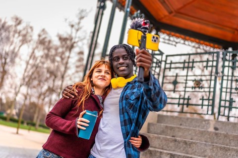 Two young content creators are recording a video blog in a park, using a smartphone attached to a