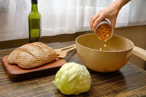 A hand pours canned corn into a mixing bowl on a wooden table. Nearby, a head of lettuce and a loaf