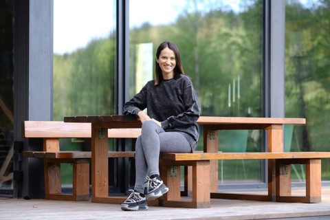 A woman with a bright smile is sitting at a rustic wooden bench on the porch of modern glass cabin,