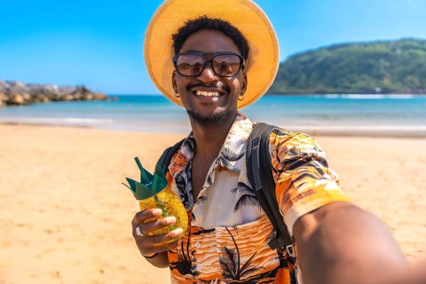 Young black man enjoying summer vacations on a tropical beach, taking a selfie while holding a