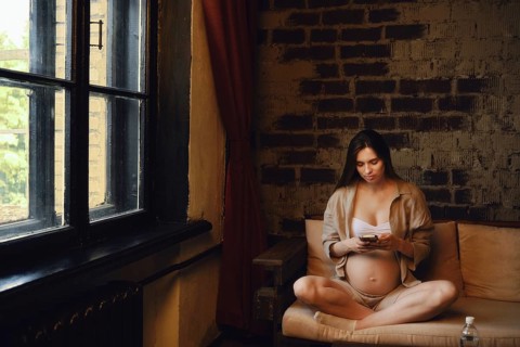 A woman in her third trimester sits comfortably on a sofa at home. She is wearing casual clothing