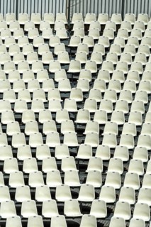 Rows of empty white seats fill a large stadium, creating a uniform pattern. The seating arrangement