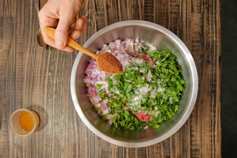 A hand holds a wooden spoon filled with spices over a stainless steel bowl containing chopped