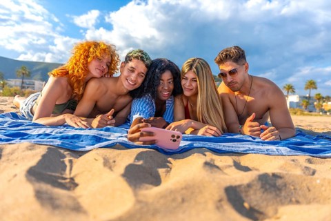 Five cheerful young friends are lying on a beach towel, taking a selfie with a smartphone, enjoying