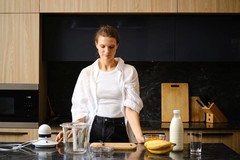 A woman stands in a contemporary kitchen, preparing to blend banana and sea buckthorn smoothies.