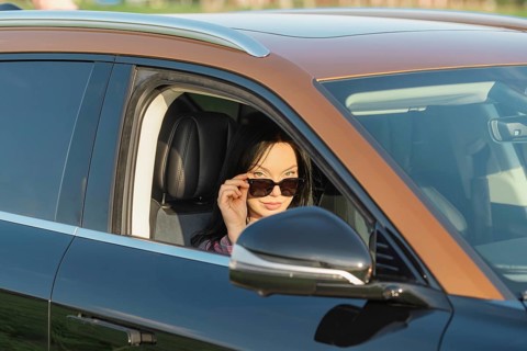 A woman with long dark hair wears stylish sunglasses while sitting in a sleek black car. The sun