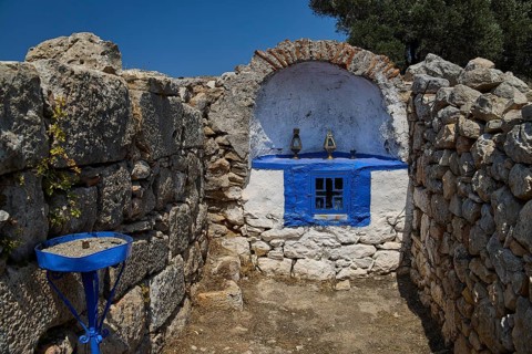 Stone chapel with blue window amidst ancient ruins, surrounded by natural walls and a deep blue