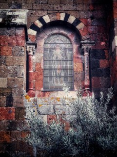 An ornate stained glass window featuring detailed artwork is set into a weathered stone wall.