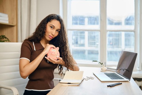 A woman with long curly hair is sitting at a desk, smiling while holding a piggy bank. She is