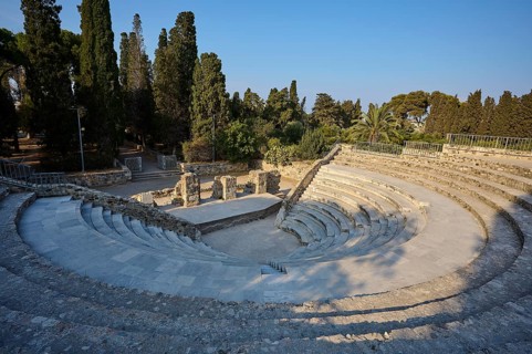Ancient round amphitheatre with stone steps, surrounded by tall trees and ruins, Ancient Theatre,