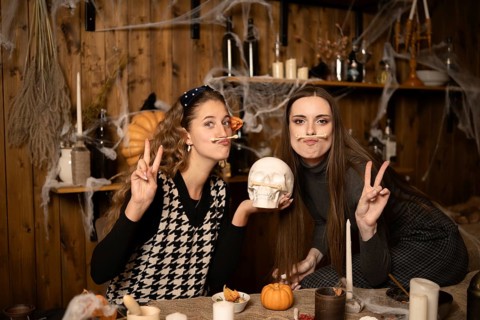 Two friends pose playfully in a decorated room for Halloween, surrounded by candles, pumpkins, and
