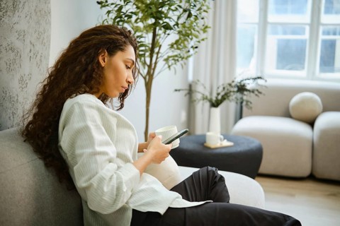 A woman sits on a comfortable sofa in a bright living room, sipping from a cup while focused on her