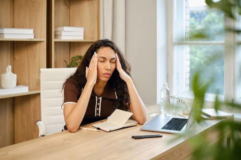 A woman with curly hair sits at a wooden desk in a stylish home office, visibly stressed while