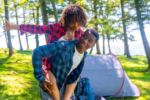 Young man giving piggyback ride to girlfriend in forest near tent, enjoying camping trip in nature