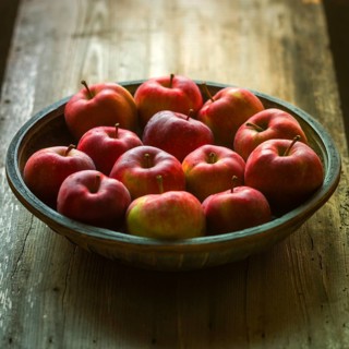 A rustic wooden bowl filled with freshly harvested red apples sits atop a weathered wooden table.