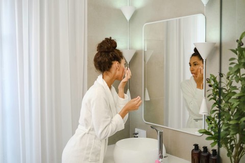 A woman in a white robe is applying skincare in a stylish bathroom. She is focused on her