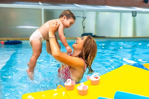 Mother playfully lifting smiling baby out of water in indoor swimming pool during matinee swim