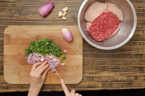 A person chops fresh herbs and onions on a wooden cutting board while ground meat rests in a