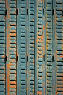A close-up view of weathered metal shutters displaying a mix of blue paint and rust. The surface