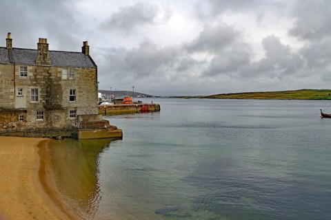 A stone cottage stands on the sandy shore of a calm loch under a cloudy sky, Lerwick, crime series,