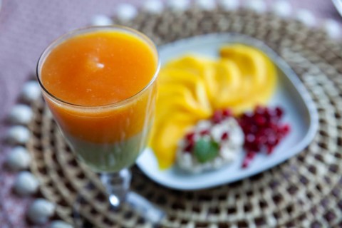 A glass with multi-coloured juice and a plate with mango slices and pomegranate seeds, Lower