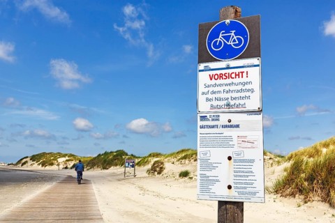 Cyclist on wooden path through the dunes to the sea, sign warns of wetness, danger of slipping and
