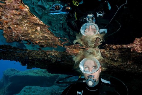 Diver with open blonde hair reflected in large air bubble in cave ceiling in underwater grotto,