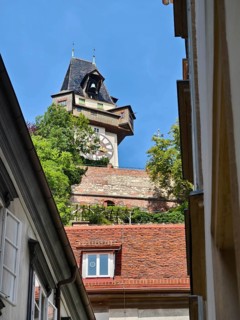 Historic clock tower towers over rooftops, surrounded by green trees and blue sky Graz Clock Tower