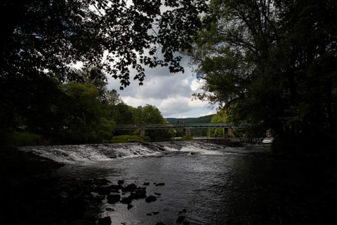 A dark river with a waterfall and bridge, surrounded by forests and cloudy skies Hardegg
