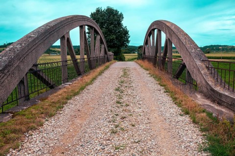An old bridge with a gravel path leads through a rural area, surrounded by fields and a clear sky
