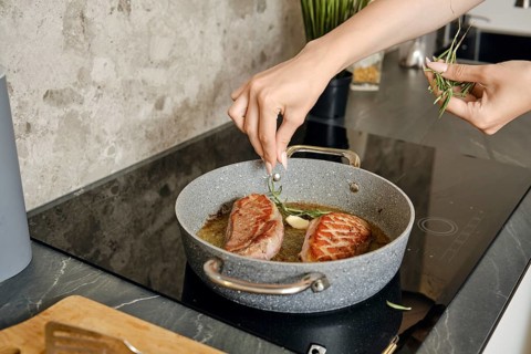 A person is preparing two pieces of duck breast in a frying pan, adding fresh herbs to enhance