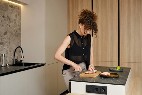 A woman with curly hair is cutting raw meat in a kitchen. Natural light illuminates her workspace