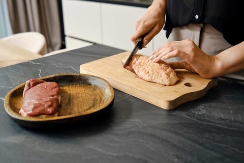 A person skillfully cuts the skin crosswise on a duck breast on a wooden board placed on a dark