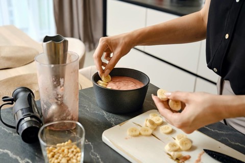 A person adds slices of bananas to a creamy smoothie mixture in a black bowl. Nearby, a blender and