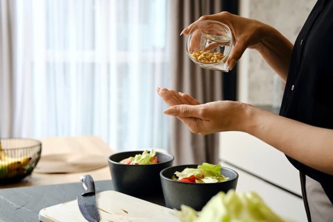 A person adds pine nuts to a bowl filled with fresh salad ingredients, including lettuce and