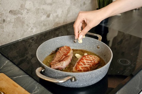 A hand adds garlic to sizzling duck steaks in a pan on the induction stovetop. The preparation