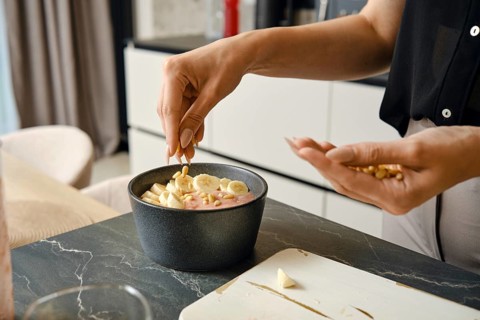 In a modern kitchen, a person adds pine nuts over sliced bananas to a bowl filled with a smoothie