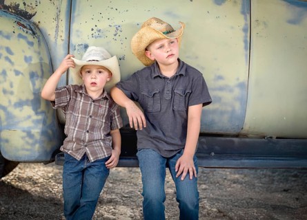Two young boys wearing cowboy hats leaning against antique truck