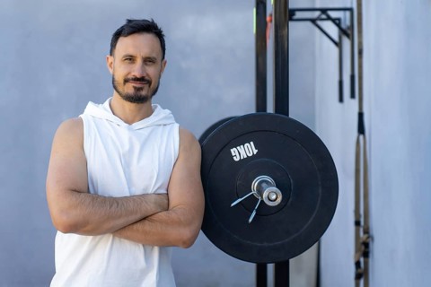 Muscular personal trainer with crossed arms leans confidently against a weight plate in an outdoor