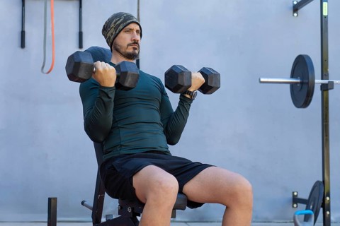 Focused male athlete sitting on a weight bench, lifting dumbbells and performing bicep curls during
