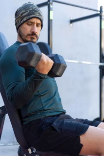 Focused athlete lifts dumbbells, performing bicep curls while seated on a bench in an outdoor gym,