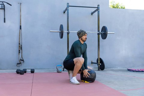 Young sportsman wearing beanie and thermal shirt performing wall ball squats in an outdoor gym,