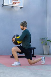 Young sportsman wearing a beanie and sportswear is lunging with a medicine ball in an outdoor gym,