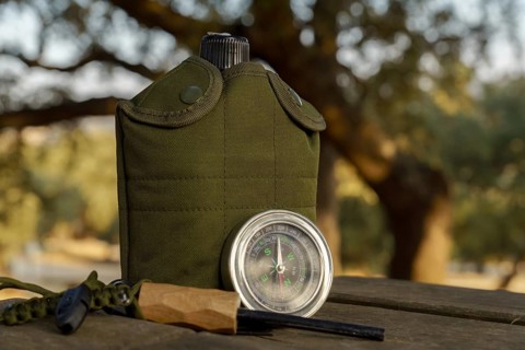 Survival gear including canteen, compass, fire starter and whistle are laying on a wooden table