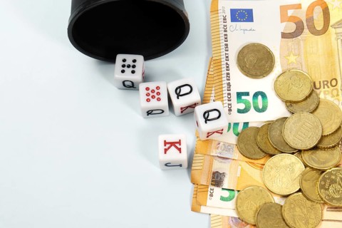 Poker dice spilling from a black cup onto a pile of fifty euro banknotes and coins, symbolizing