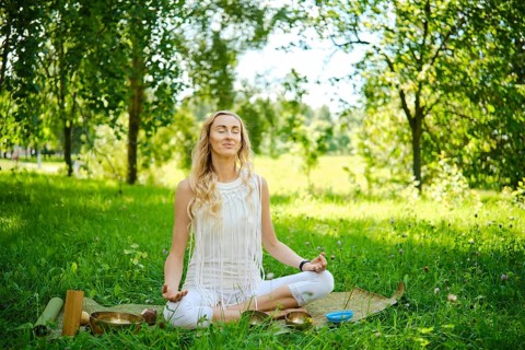 A woman sits cross-legged on a mat in a lush green park, eyes closed and arms resting on her knees.