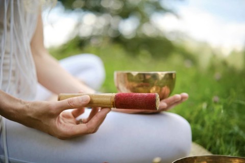 A person is holding a red-tipped mallet next to a singing bowl, seated on grass in a serene outdoor