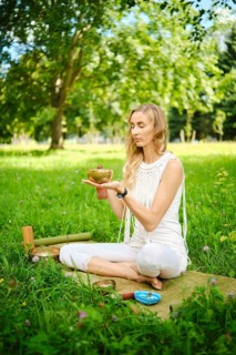 A woman sits cross-legged in a grassy park, holding a singing bowl while meditating. Surrounded by