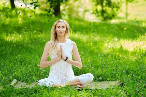 A woman dressed in white sits cross-legged on a mat, practicing yoga outdoors. Surrounded by