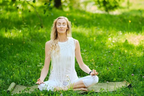 A woman in a white outfit sits cross-legged on a mat, eyes closed and hands resting on her knees in